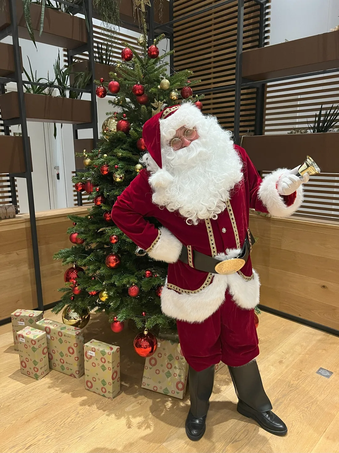 Le Père Noël en costume complet devant un sapin décoré.